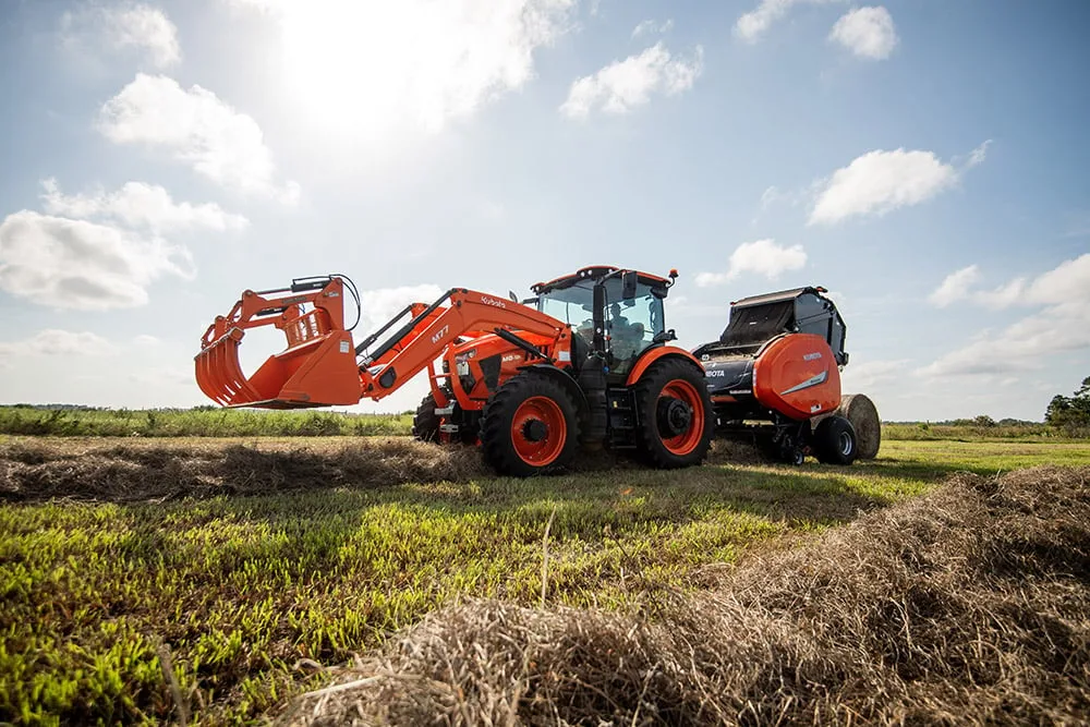 Kubota M8-181 tractor with a loader grapple beside a Kubota round baler in a hay field