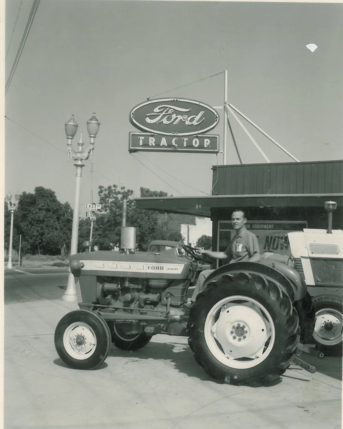 Man seated on a Ford 3000 tractor on the sidewalk beneath the neon Ford Tractor sign