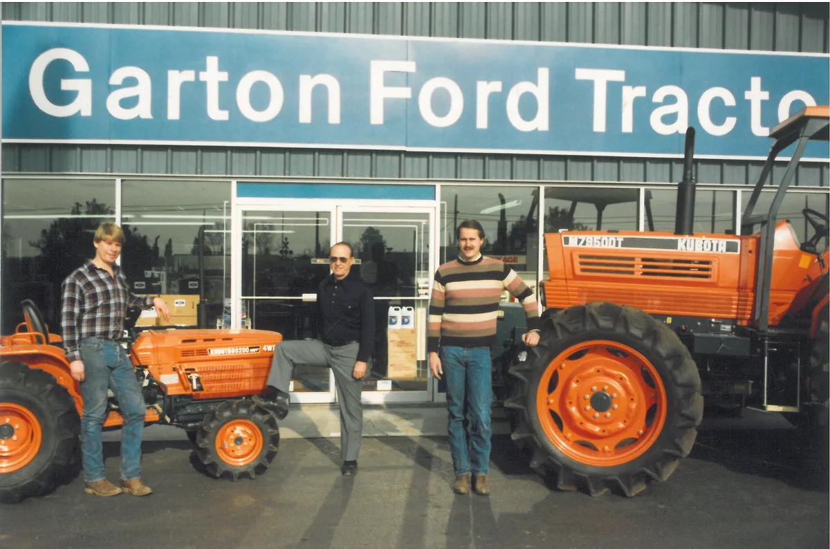 Three men with two orange Kubota tractors in front of the Garton Ford Tractor storefront