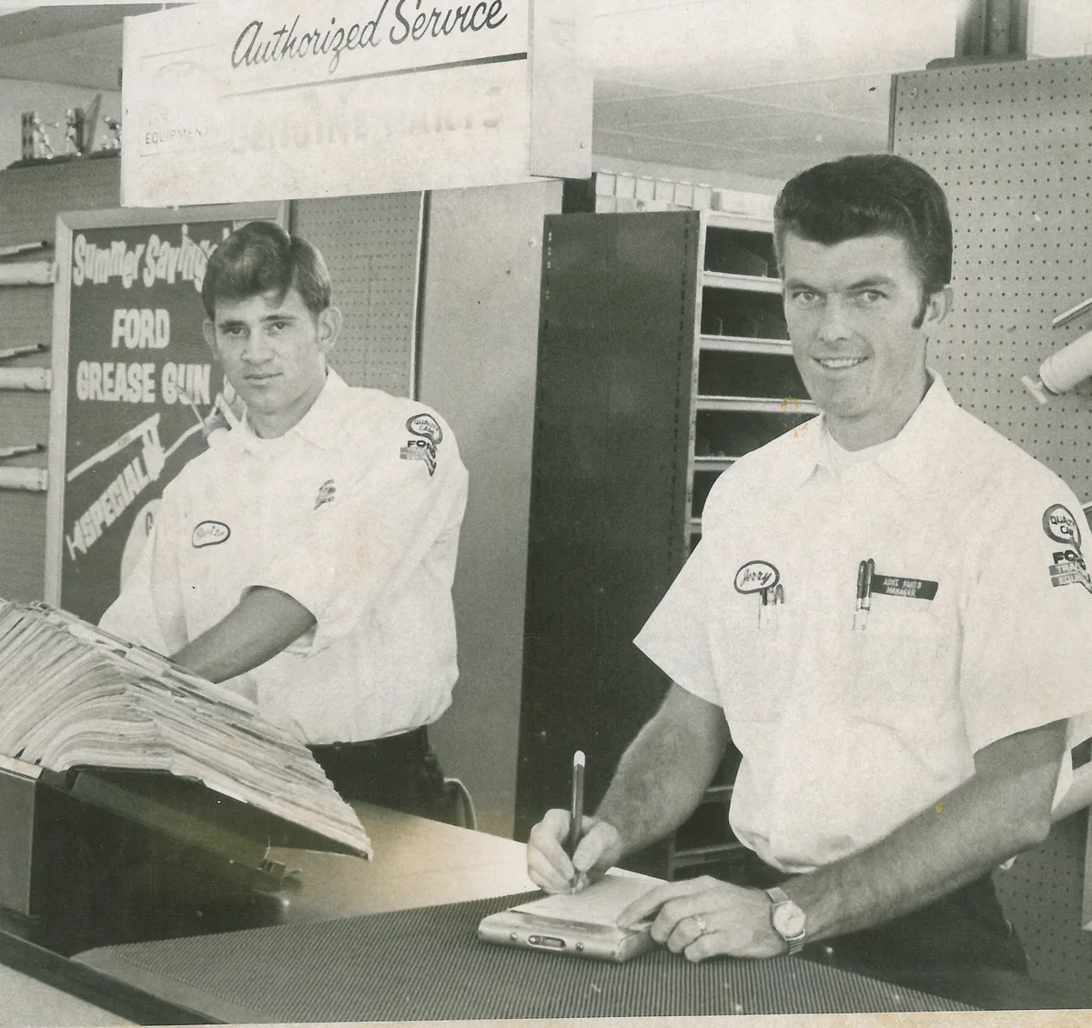 Two parts employees in white uniform shirts behind the parts counter, one writing an order, beneath an Authorized Service sign