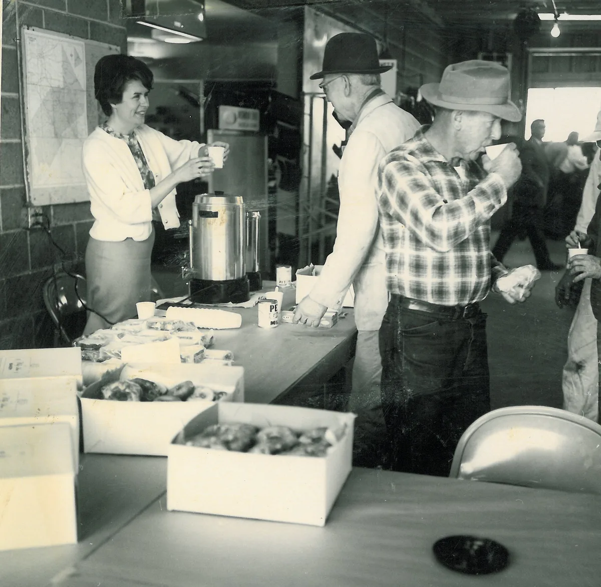 Woman pouring coffee from an urn for customers beside boxes of doughnuts at a table inside the shop