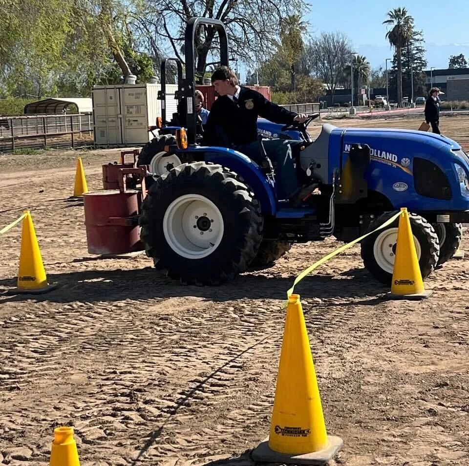 A student in an FFA jacket operating a blue New Holland tractor between yellow cones